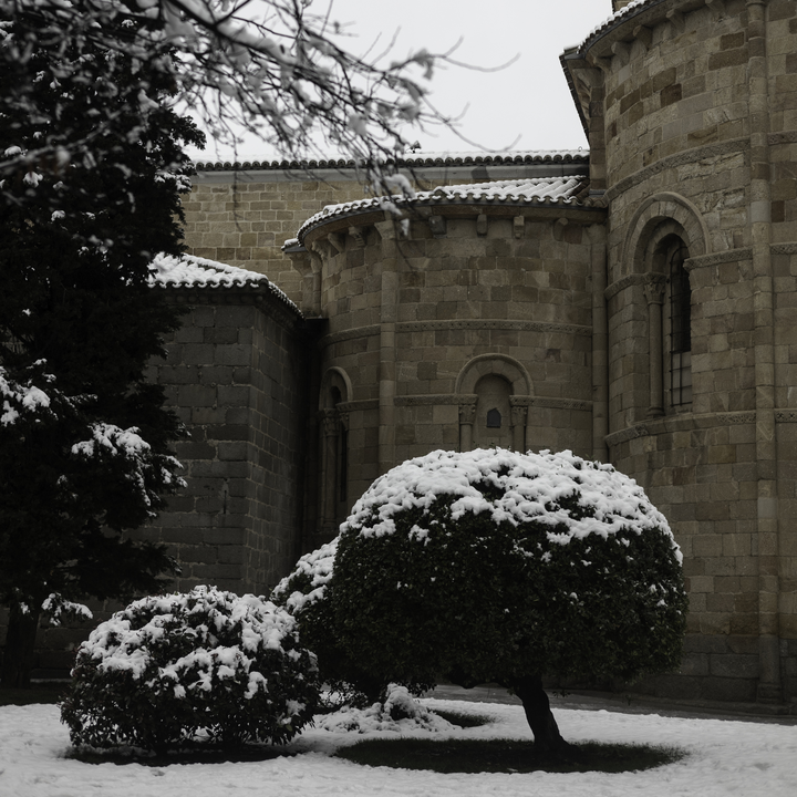 Heavy snow blankets the garden bushes outside the rear Romanesque exterior of San Pedro Church in Avila, Spain, highlighting the ancient stone architecture on a freezing winter day.