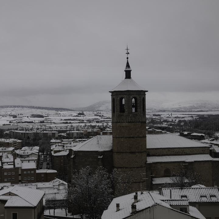 An elevated winter view overlooking the snowy Church of Santiago the Apostle and residential rooftops in southern Avila, Spain, with the snow-capped Gredos Mountains visible in the distance under an overcast sky.