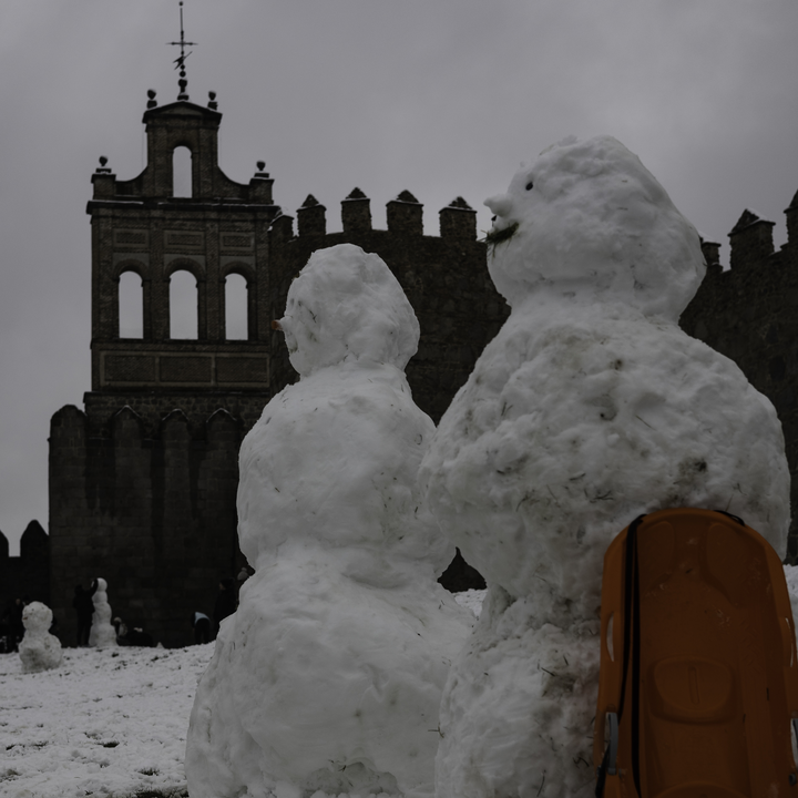A lively winter scene at the northern stone fortifications of Avila, Spain, where locals and visitors play in the deep snow and build snowmen against the backdrop of the historic medieval walls.