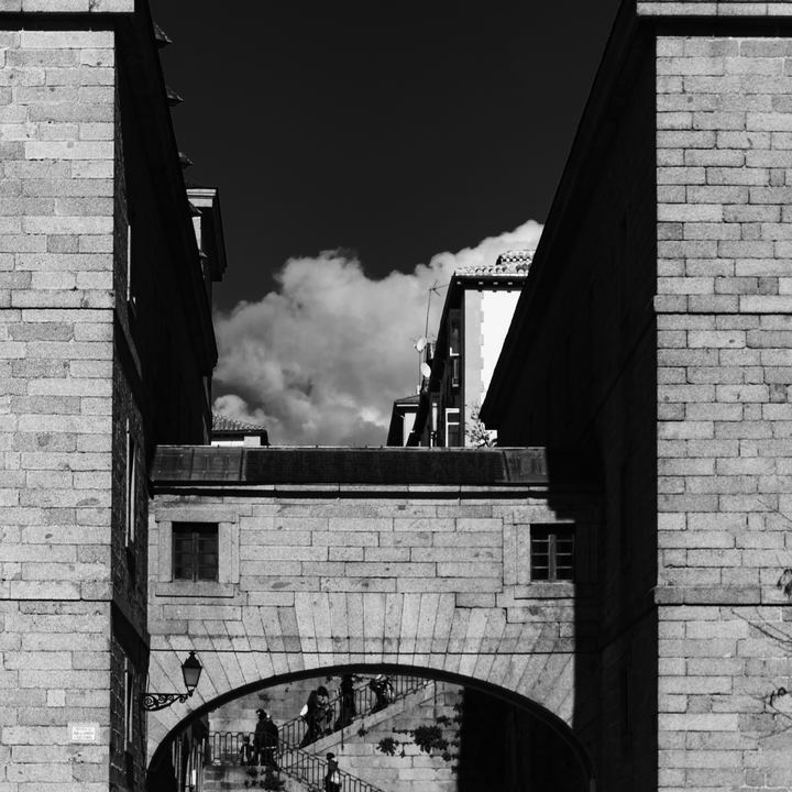  Capturing the architectural charm of San Lorenzo de El Escorial, featuring a stone archway that connects two imposing stone buildings. Above the arch sits a bridge-like structure with a small window, while below, people ascend and descend a staircase that leads through the archway. The interplay of historic textures, light, and shadow contrasts beautifully with modern buildings visible in the background, highlighting Spain's cultural layers and timeless urban design. 