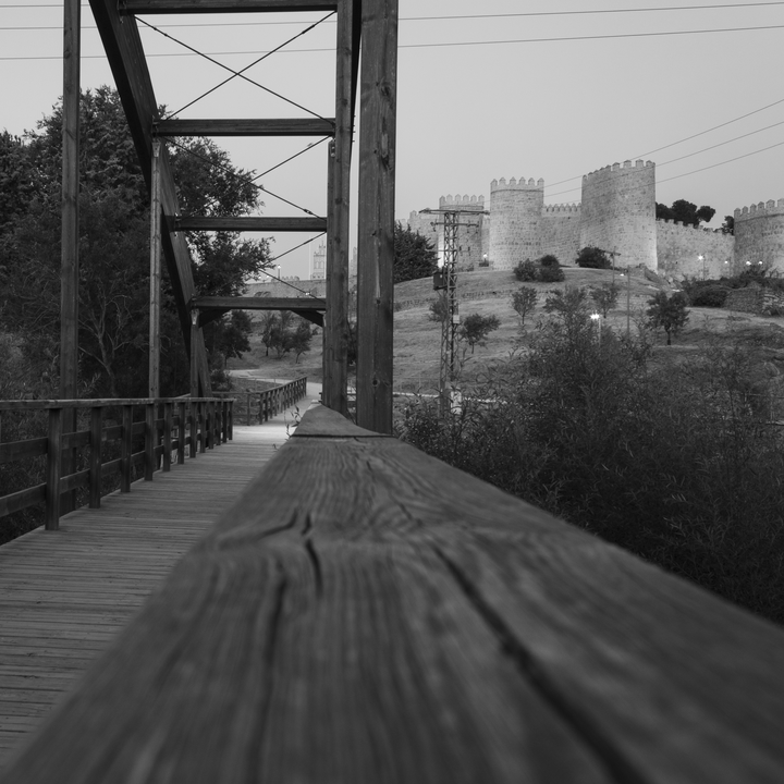 Striking black and white photograph captures the approach to Ávila's iconic medieval stone wall through a rustic wooden bridge with railings and overhead metal cross-bracing. The scene blends natural surroundings—trees and bushes—with historical architecture featuring rounded towers and fortification elements. The foreground emphasis on the bridge's railing enhances depth and perspective, creating a dramatic visual transition between nature and centuries-old Spanish heritage. 