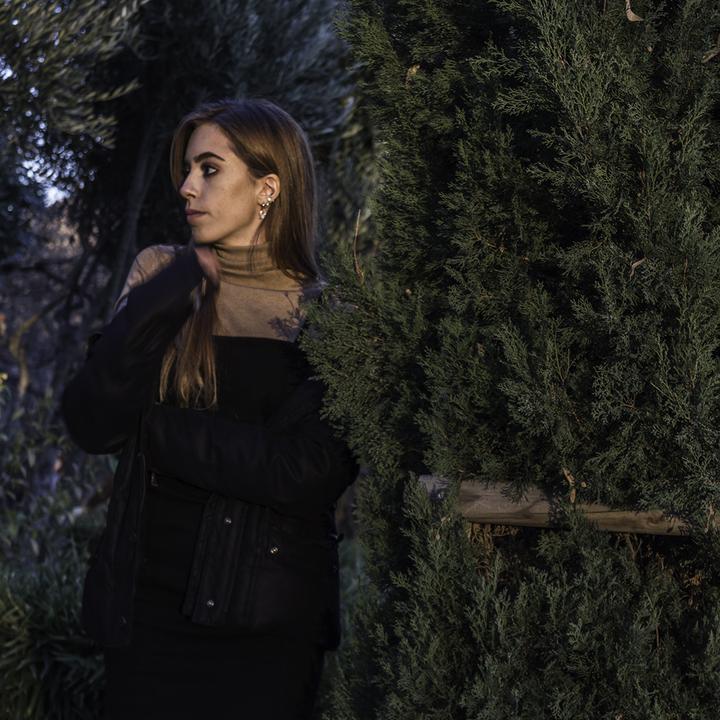 Woman in Black Jacket and Dress Standing Near Green Bush in Outdoor Setting. 