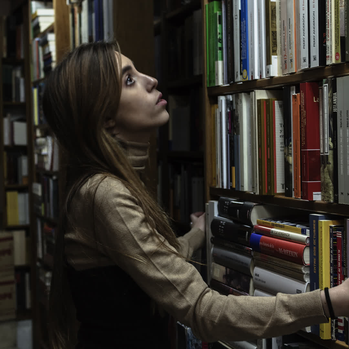 A young woman stands thoughtfully in a quaint bookshop, browsing through shelves filled with books. The warm and quiet atmosphere invites exploration and discovery. 