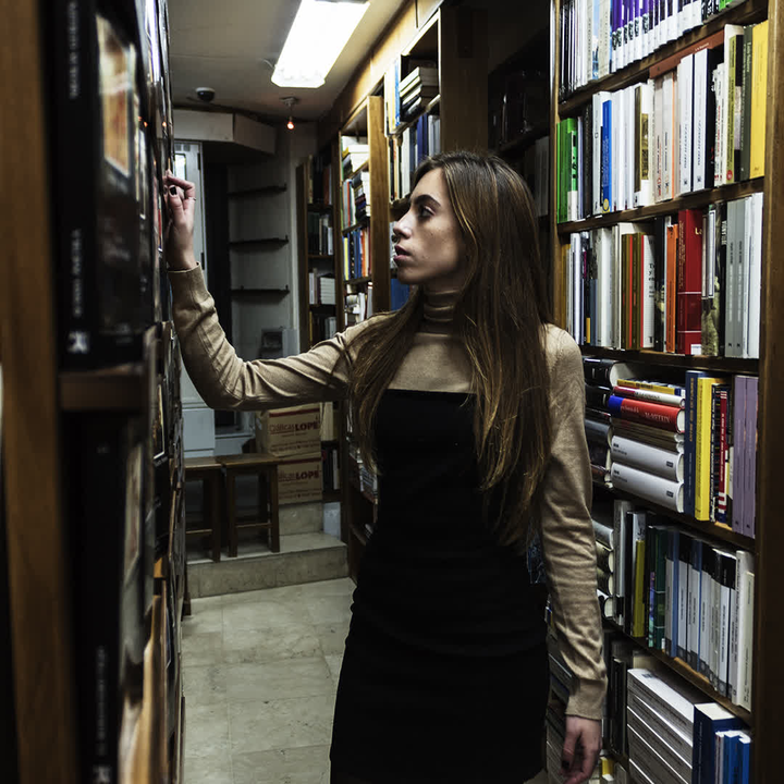 A young woman stands thoughtfully in a quaint bookshop, browsing through shelves filled with books. The warm and quiet atmosphere invites exploration and discovery. 