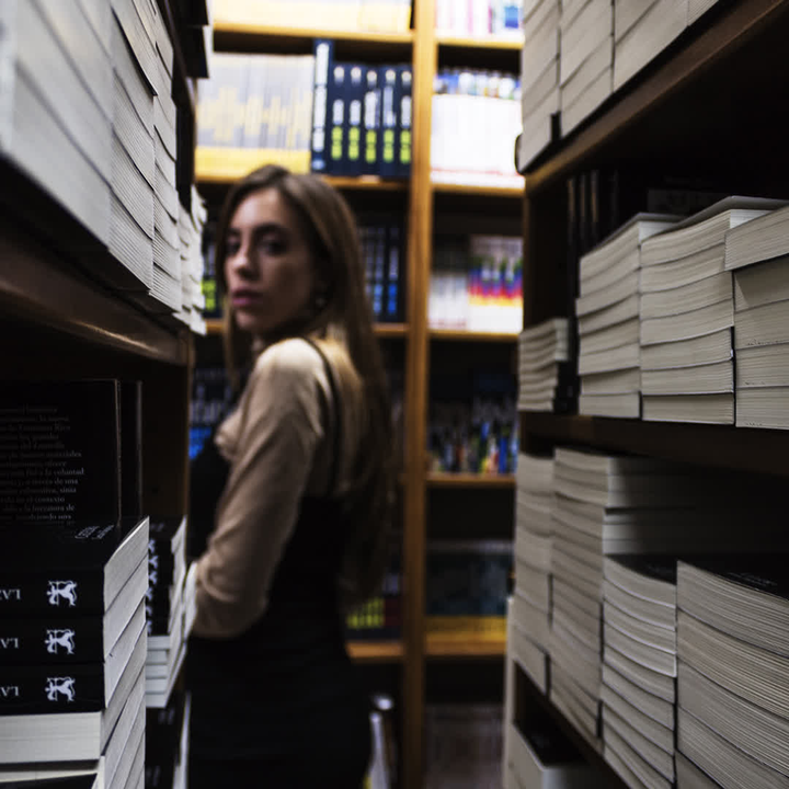 A young woman stands thoughtfully in a quaint bookshop, browsing through shelves filled with books. The warm and quiet atmosphere invites exploration and discovery. 