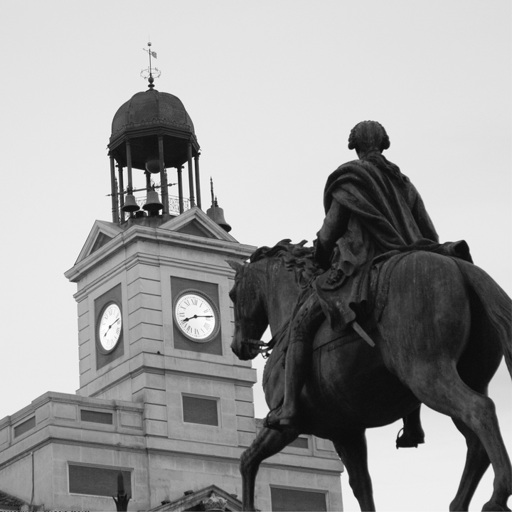  Black and white photograph of the famous Puerta del Sol in Madrid, capturing the equestrian statue of King Charles III and the historic clock tower of the Real Casa de Correos, a central symbol of the city. 