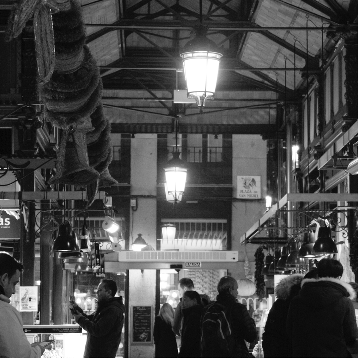  Black and white photograph of the bustling interior of Madrid's Mercado de San Miguel, highlighting the elegant ironwork, hanging cured meats, warm lighting, and lively ambience of this iconic food market. 