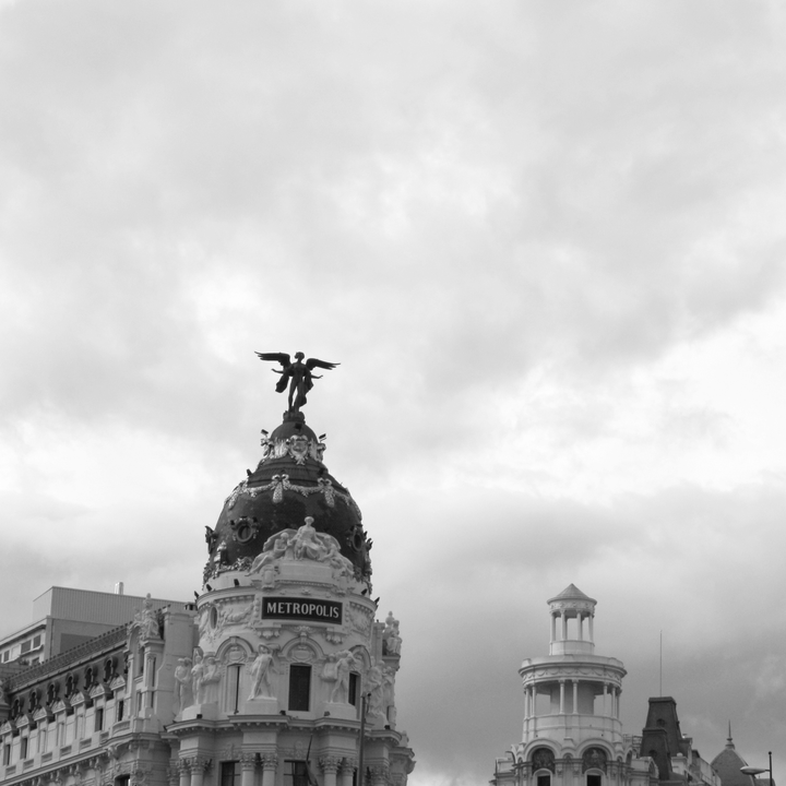 Black and white image of the iconic Metropolis Building on Gran Vía in Madrid, Spain, featuring ornate Beaux-Arts architecture with the famous winged statue against a moody sky backdrop. 
