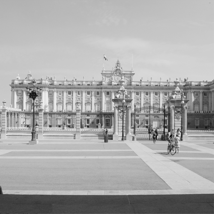  A timeless black and white photograph of the Royal Palace of Madrid (Palacio Real), capturing the grandeur of the architecture under a clear sky with pedestrians and cyclists animating the forecourt. 
