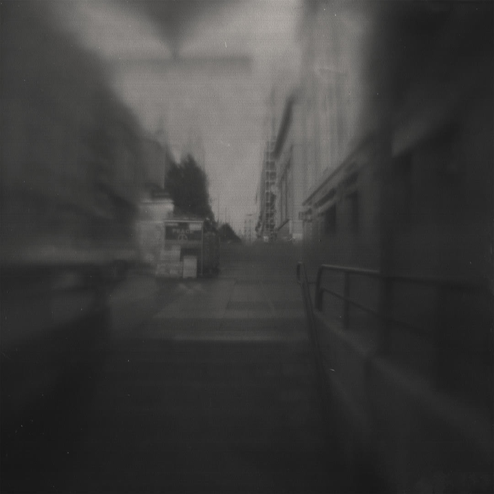 A blurred black-and-white photograph capturing an urban scene near Metro Gran Vía in Madrid. The image evokes a sense of motion and nostalgia, with soft tones and abstract details enhancing its atmospheric quality. Camera design by @joterman. 