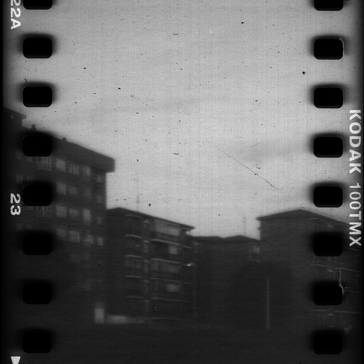 Analog black and white street photography of an urban scene in Avila, captured on Kodak 100TMX film reel. Featuring street lamps and buildings, this vintage film aesthetic evokes nostalgia and timeless urban imagery