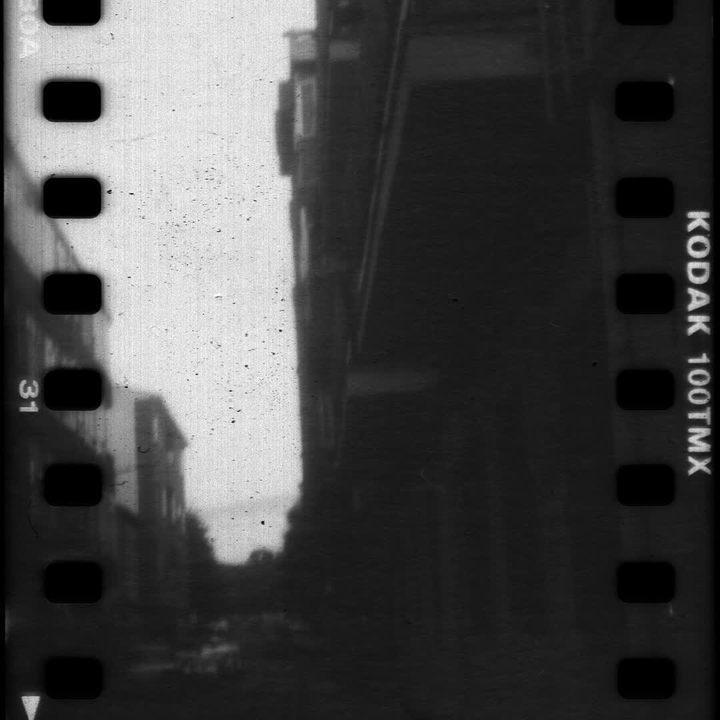 Analog black and white street photography of an urban scene in Avila, captured on Kodak 100TMX film reel. Featuring street lamps and buildings, this vintage film aesthetic evokes nostalgia and timeless urban imagery